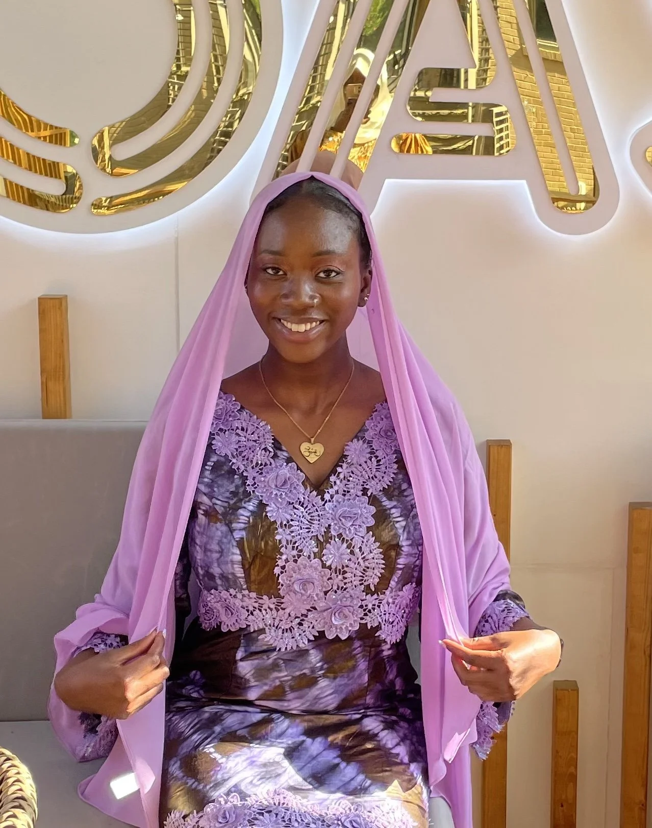 A smiling woman wearing a purple lace dress and matching headscarf, sitting indoors with a decorative wall behind her.