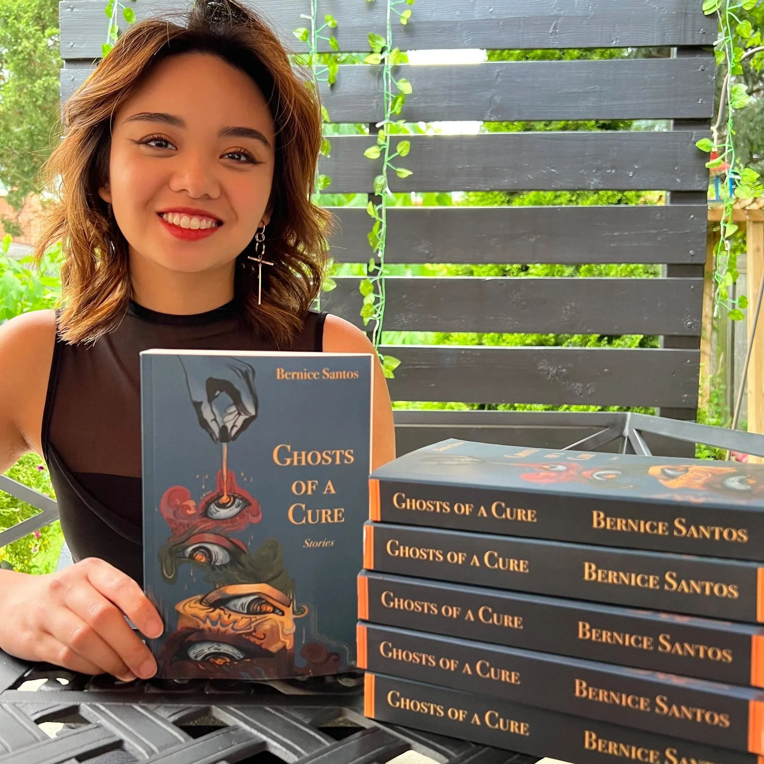 Bernice Santos, Canadian writer, editor and copywriter, holding her book Ghosts of a Cure beside a stack of copies.