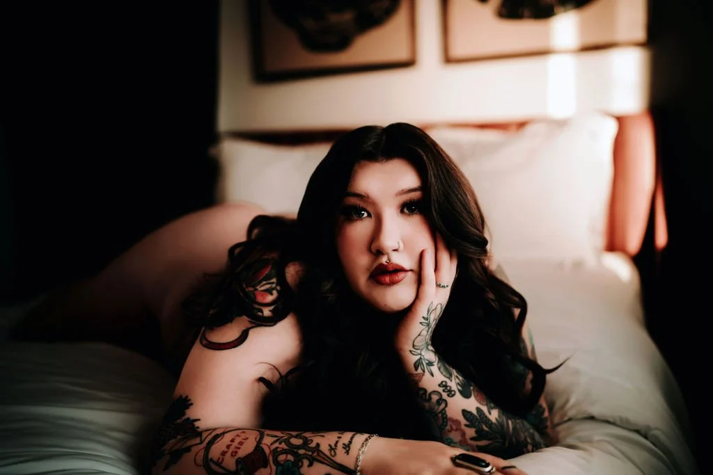 A woman with dark hair and tattoos lying on a bed, resting her face on her hand, in a dimly lit room.