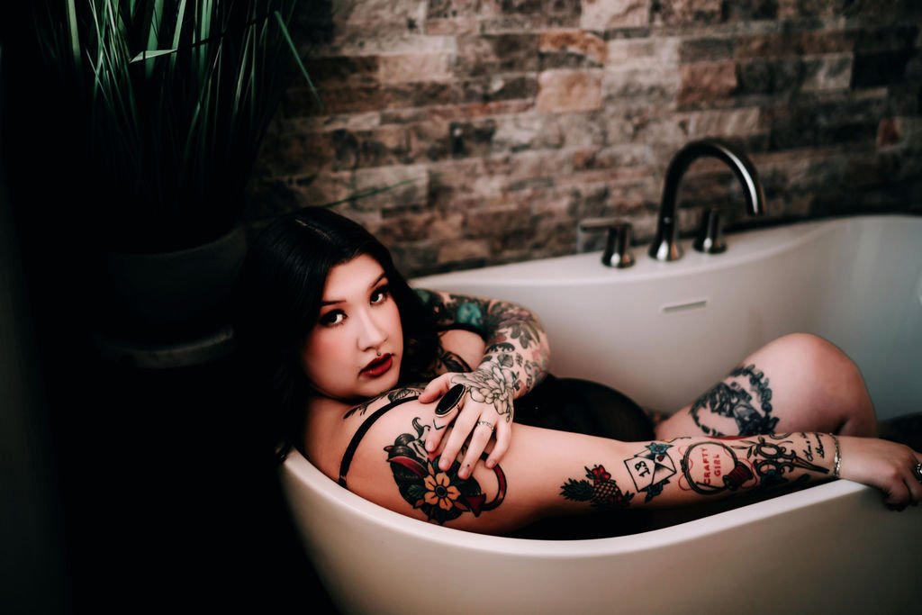 A woman with black hair and tattoos relaxing in a bathtub against a brick wall background.
