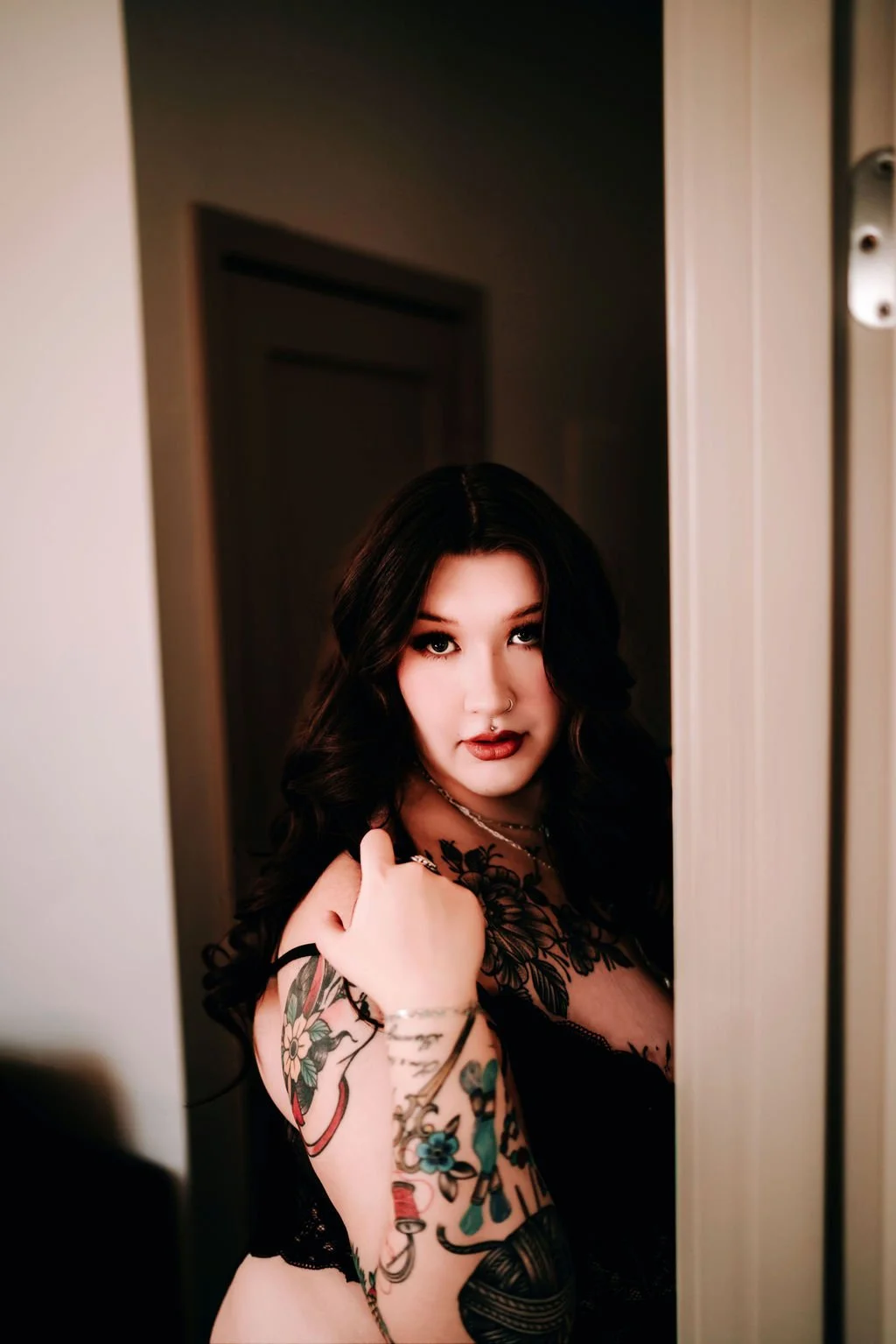 A young woman with dark, curly hair and tattoos on her arm is looking into a mirror, wearing dark lipstick and jewelry, with a dimly lit background.