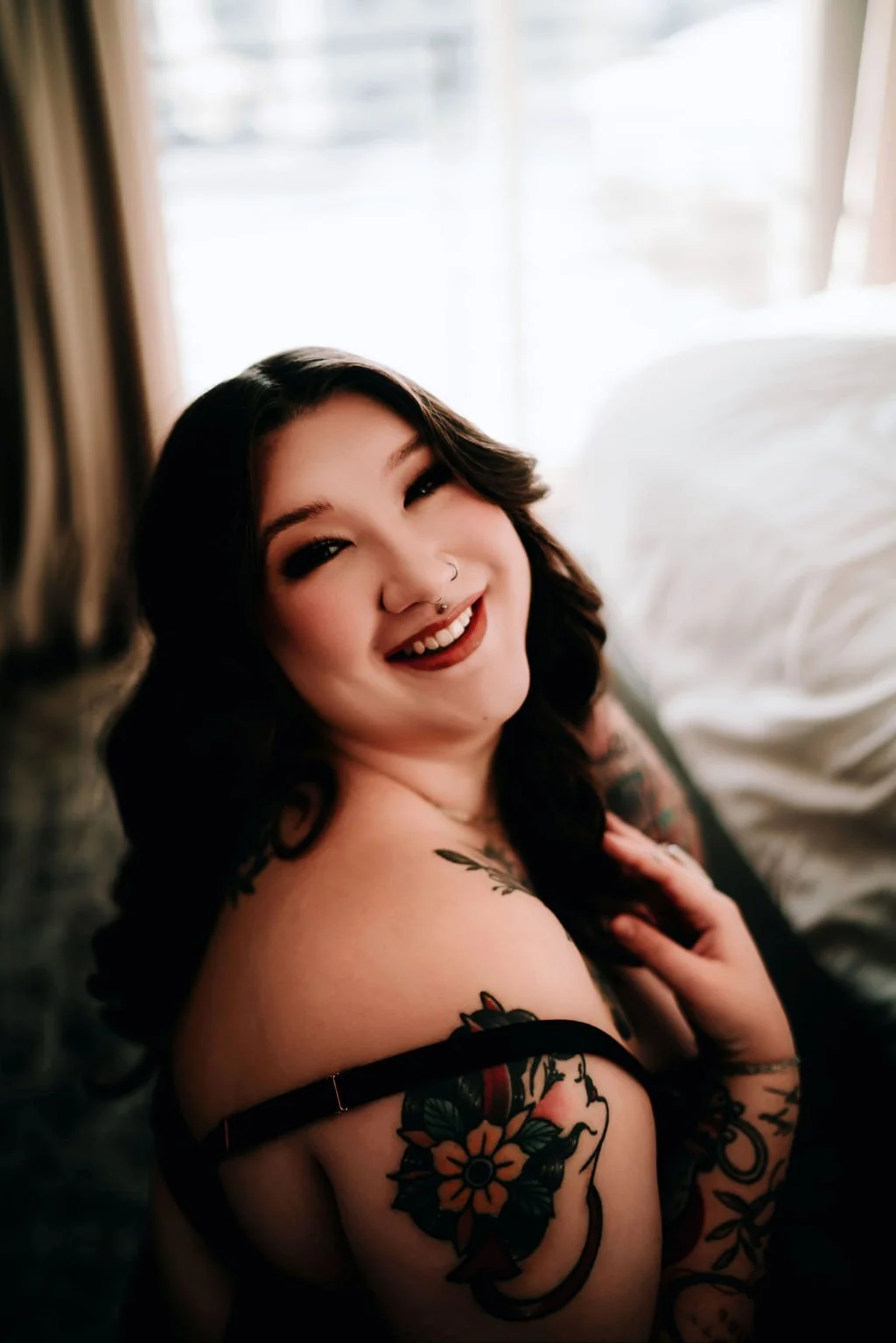A smiling woman with dark wavy hair and tattoos on her shoulder and arm, sitting near a window with sunlight, wearing a black strap top.