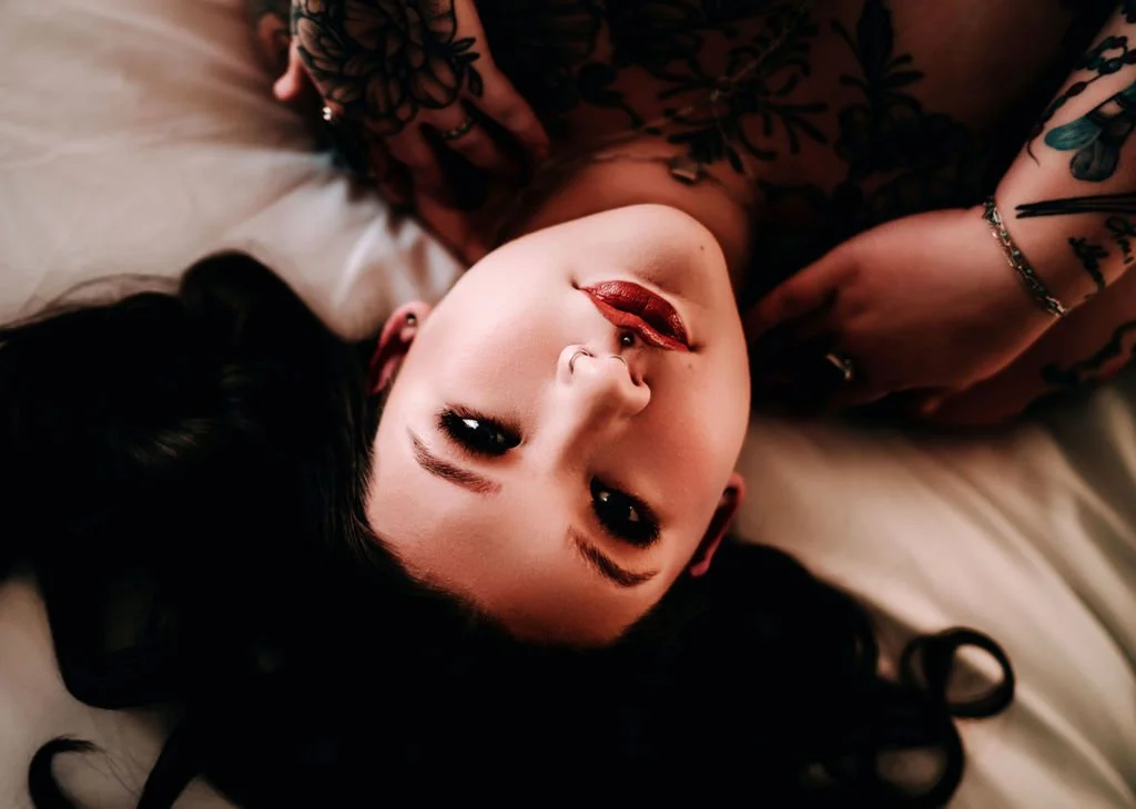 A woman lying on a bed, looking up at the camera with dark hair, red lipstick, and tattoos on her arms.