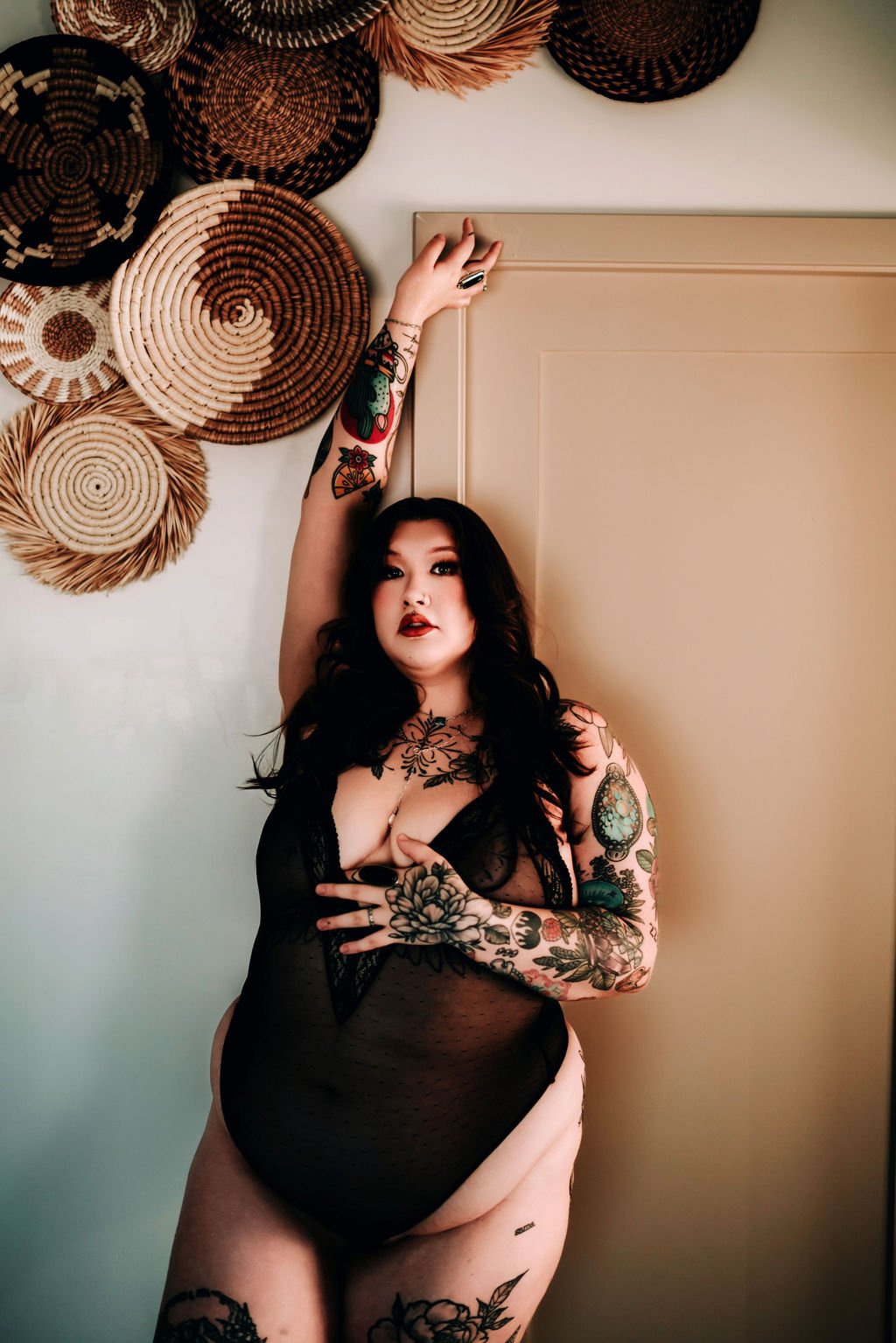 A woman with dark hair and tattoos wearing black lace lingerie, striking a pose indoors near a door, with woven baskets on the wall behind her.