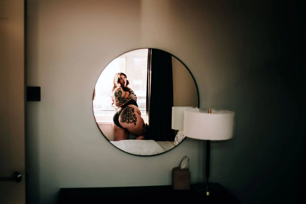A woman with tattoos takes a selfie in a round mirror on a wall, showing her reflection and part of a bed and window behind her.
