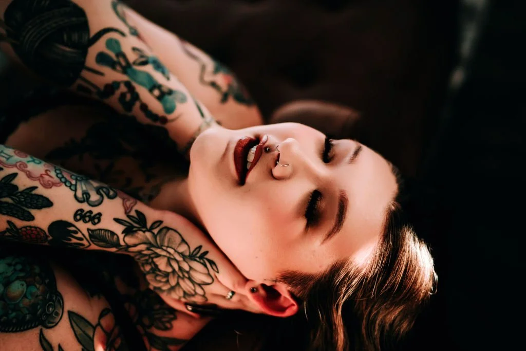 A woman with tattoos and a nose piercing lying on her back, smiling with eyes closed.