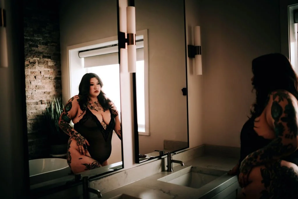 A woman with long dark hair, tattoos on her arms and legs, wearing a black lingerie bodysuit, looking at her reflection in a bathroom mirror.