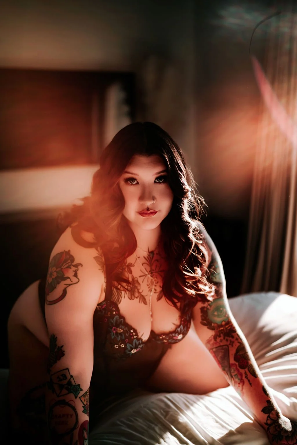 A woman with long dark hair, tattoos, and makeup, sitting on a bed in a dimly lit room with sunlight coming through curtains.