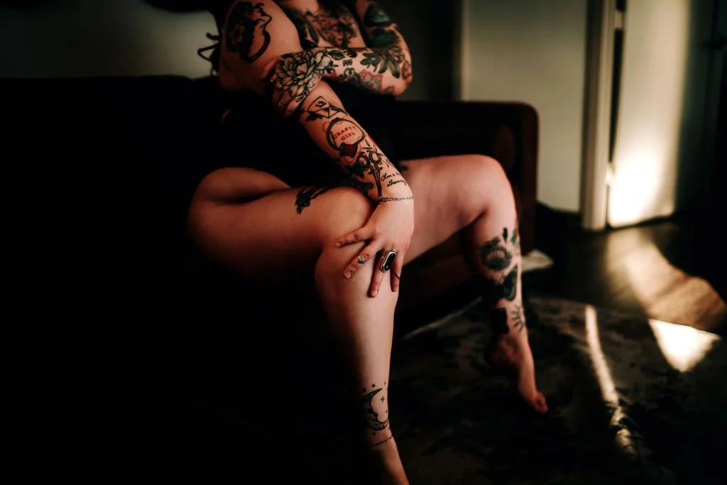 A person sitting on a dark sofa with their legs crossed, showing multiple tattoos on their arms and legs, in a dimly lit room with warm lighting.