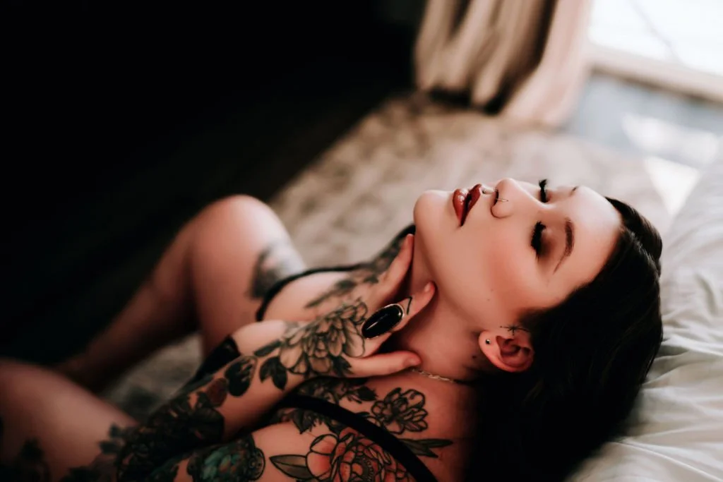 A woman with tattoos and black hair lying on a bed with her eyes closed and head tilted back.