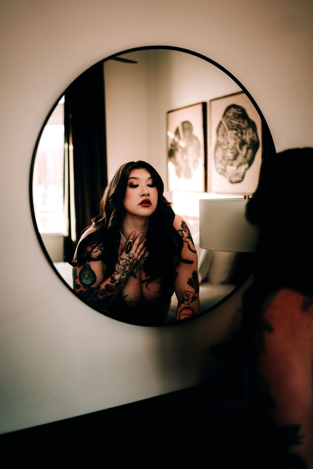 A woman with dark hair and tattoos looking at her reflection in a round mirror, with an art print on the wall behind her.