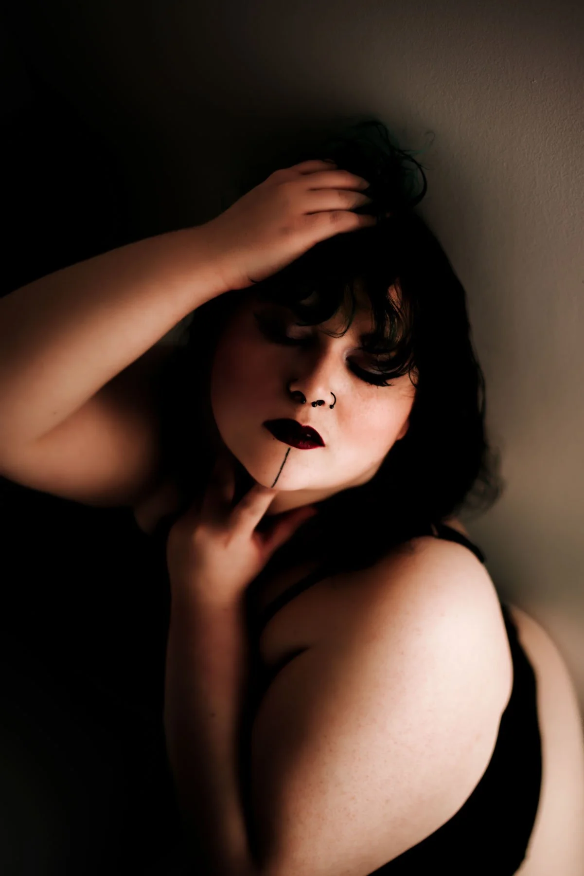 A woman with dark curly hair, dark makeup, and a septum piercing, posing with one hand on her head and the other on her chin, wearing a black strap top.