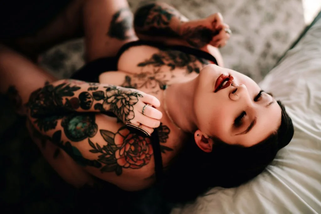 A woman with dark hair and tattoos lying on a bed, smiling with closed eyes.