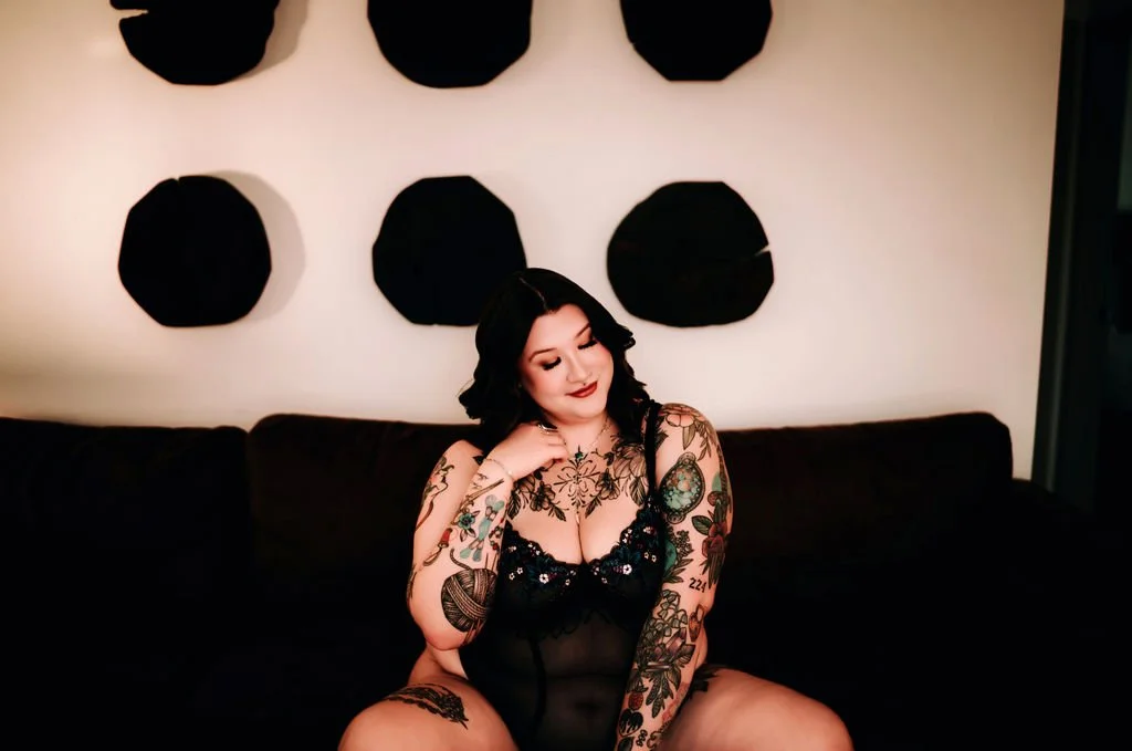 A woman with dark hair and tattoos, wearing black lingerie, sitting on a black couch in a room decorated with black art pieces on the wall.