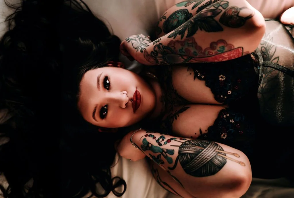 A woman lying on her back with dark hair spread around her, looking up at the camera, with prominent tattoos on her arms and chest, wearing a black lace top.