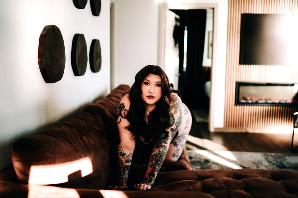 A woman with long dark hair and tattoos on her arms and legs sitting on a brown couch in a modern living room.