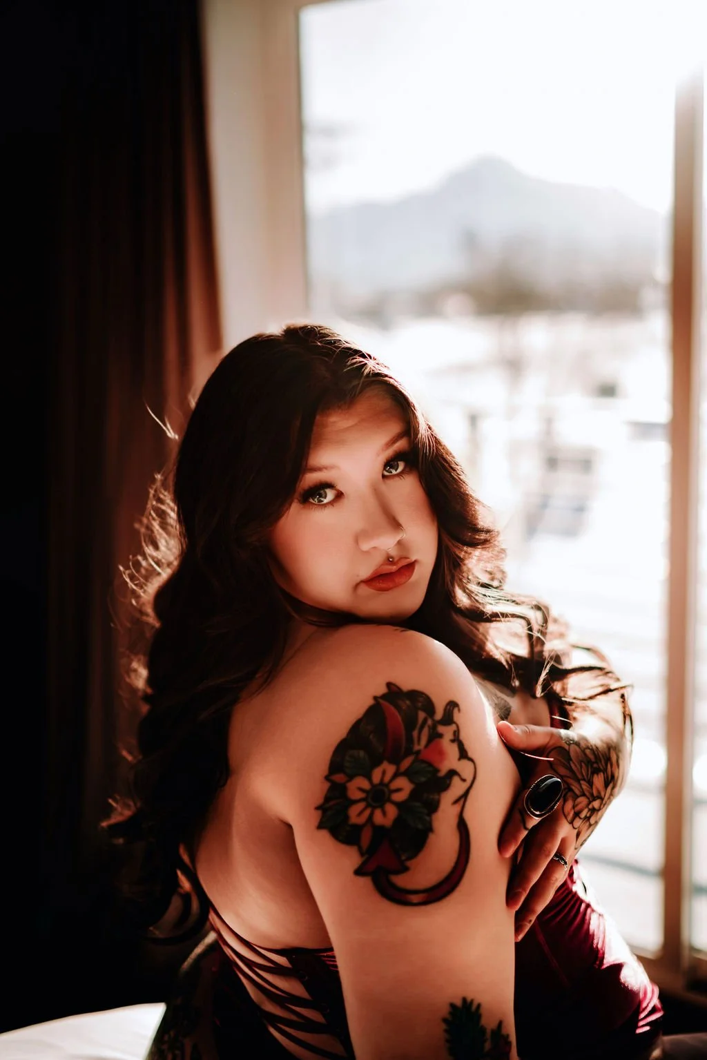 A woman with dark wavy hair looking over her shoulder at the camera inside a room with sunlight streaming through a window.