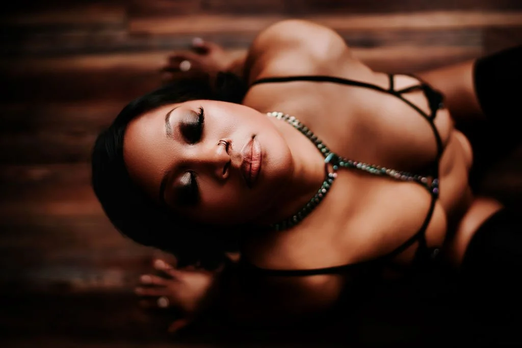 A woman with dark hair and makeup lying on a wooden floor, dressed in black lingerie and a beaded necklace.