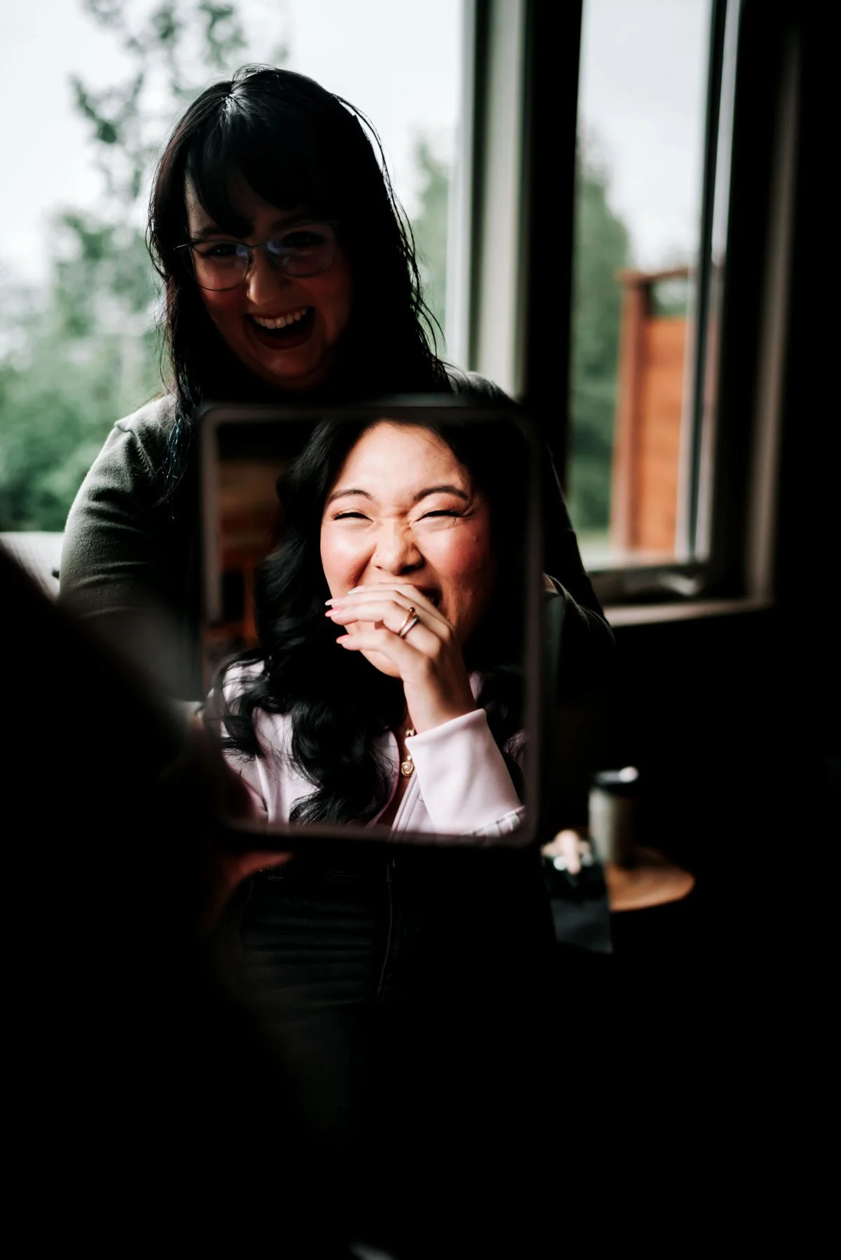 Two women are laughing and smiling while taking a photo of each other with a smartphone in front of a mirror, in a room with large windows and greenery outside.