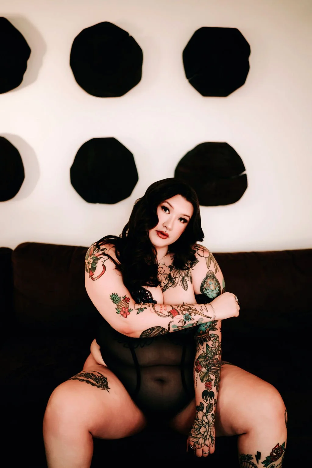 A tattooed woman with black hair and makeup, wearing black lingerie, sitting on a black couch in front of a white wall with black geometric wall decor.