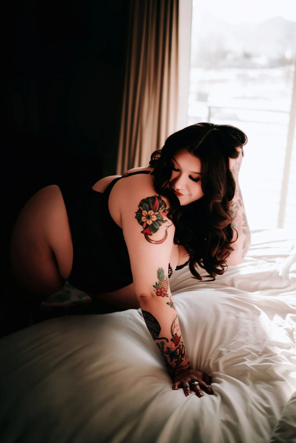 A woman with dark hair and tattoos poses on a bed, wearing black lingerie, with a window and curtains in the background.
