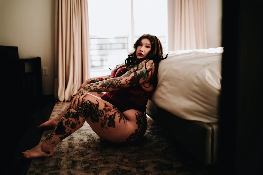 A woman with tattoos sitting on the floor by a bed in a hotel room, looking at the camera with a serious expression.