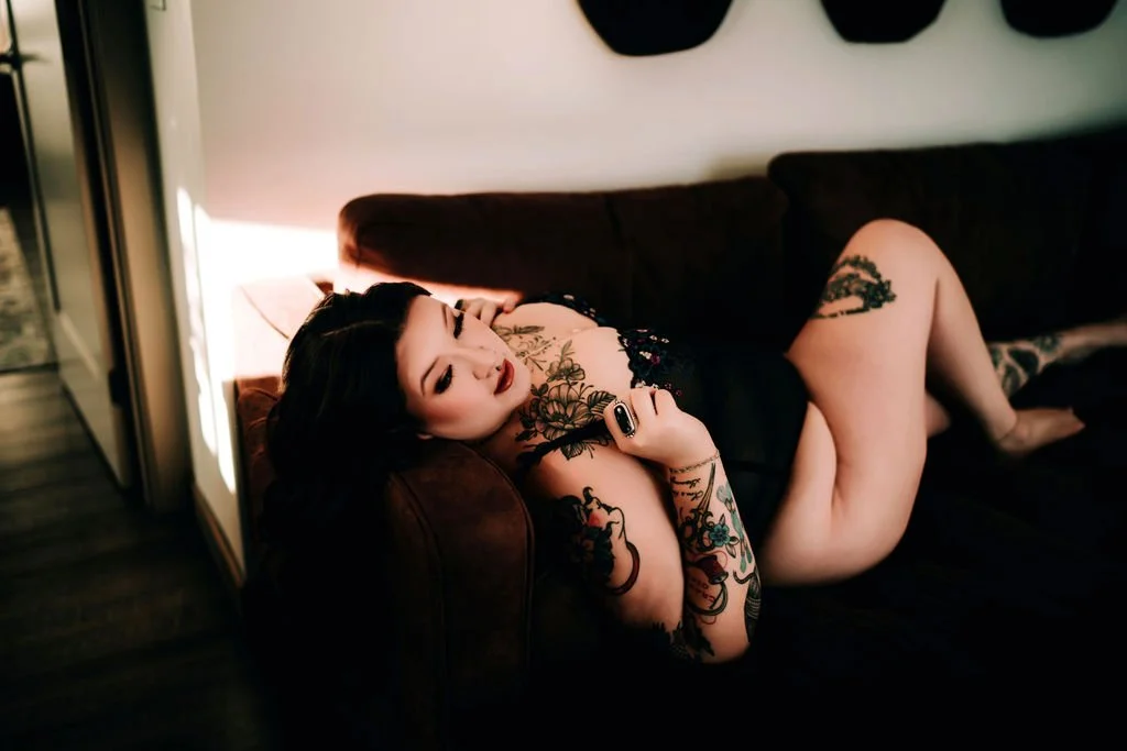 A woman with dark hair and tattoos lying on a dark couch, wearing a black lace lingerie, holding a remote control, in a dimly lit room.