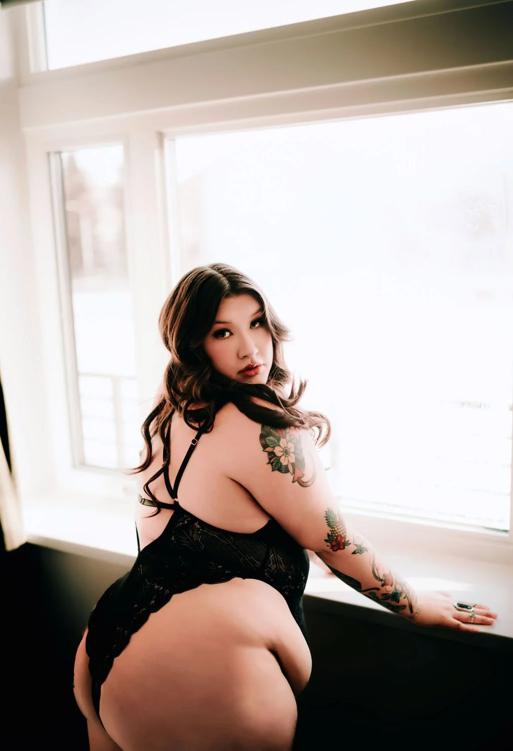 A woman with dark, wavy hair and tattoos on her arm, wearing black lingerie, leaning on a windowsill and looking at the camera with a serious expression.