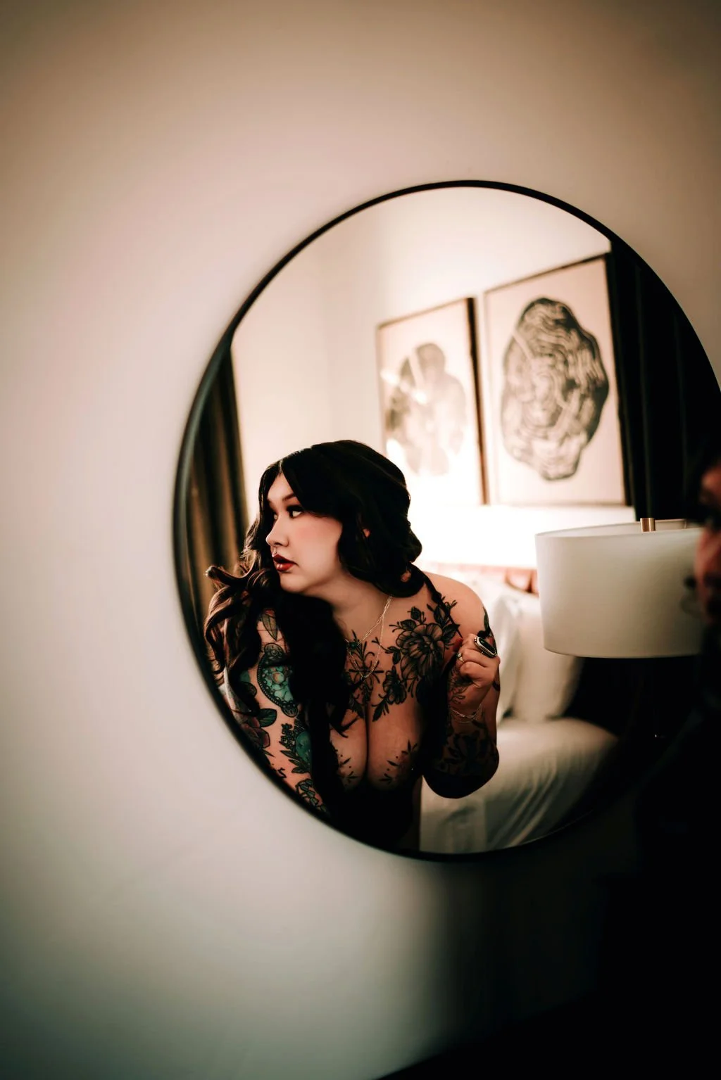 A woman with long dark hair and tattoos looking into a round mirror.