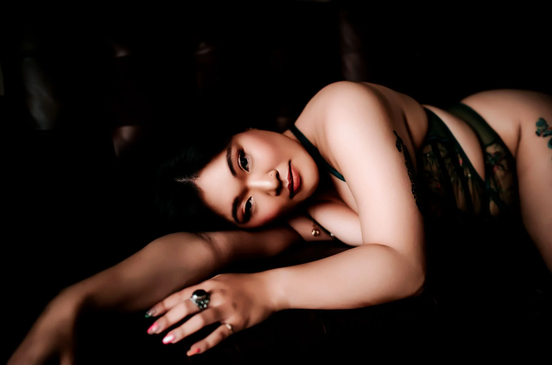 A woman with dark hair and makeup lying on a dark surface, resting her head on her arm, wearing black lace lingerie and jewelry.