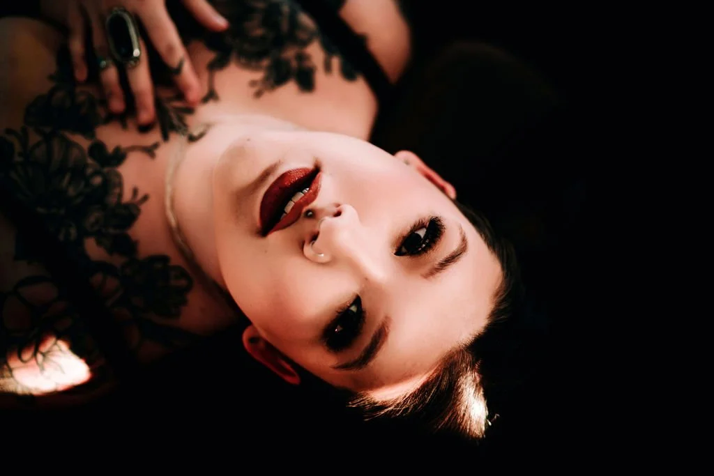 A woman with dark hair lying on her back, looking up at the camera, with makeup and a black lace top