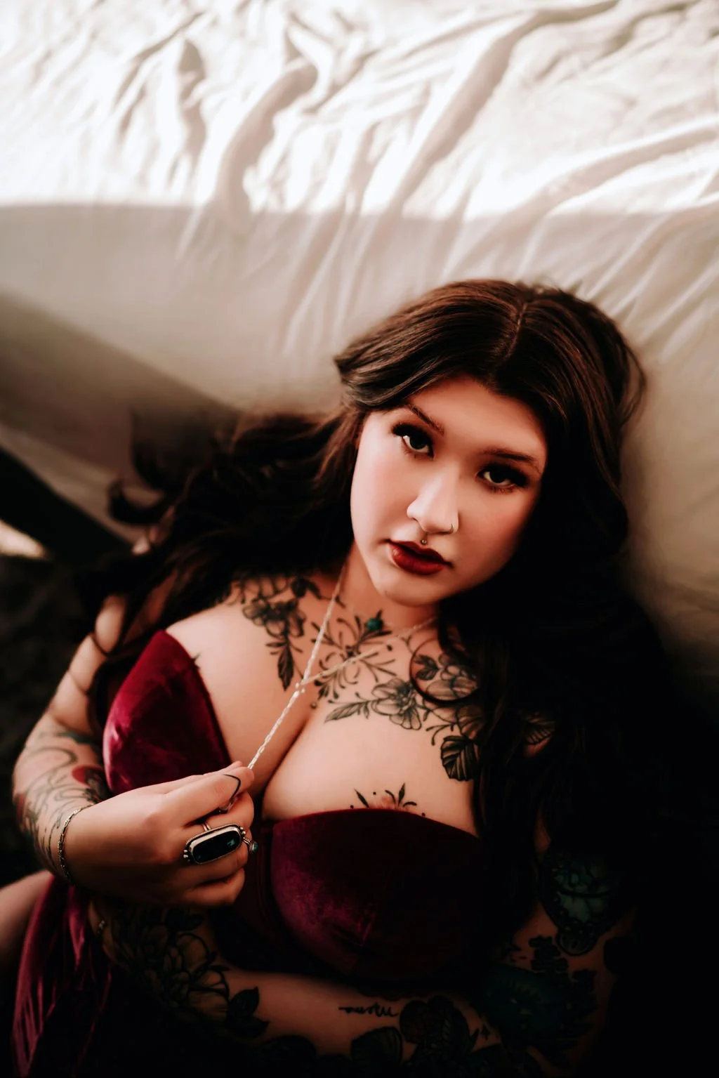 A woman with long dark hair and tattoos lying on a bed, wearing a burgundy top with a deep neckline, holding a necklace, and looking at the camera.
