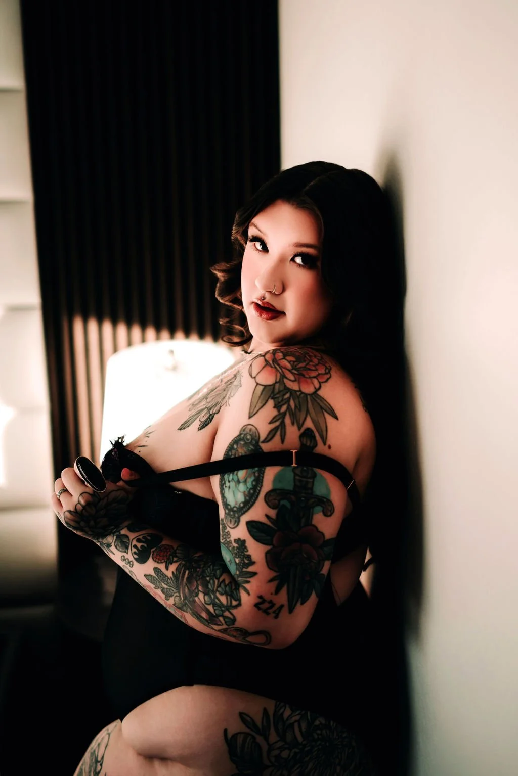 A woman with dark hair and tattoos on her arms and shoulders, wearing black lingerie, leaning against a wall in a dimly lit room.