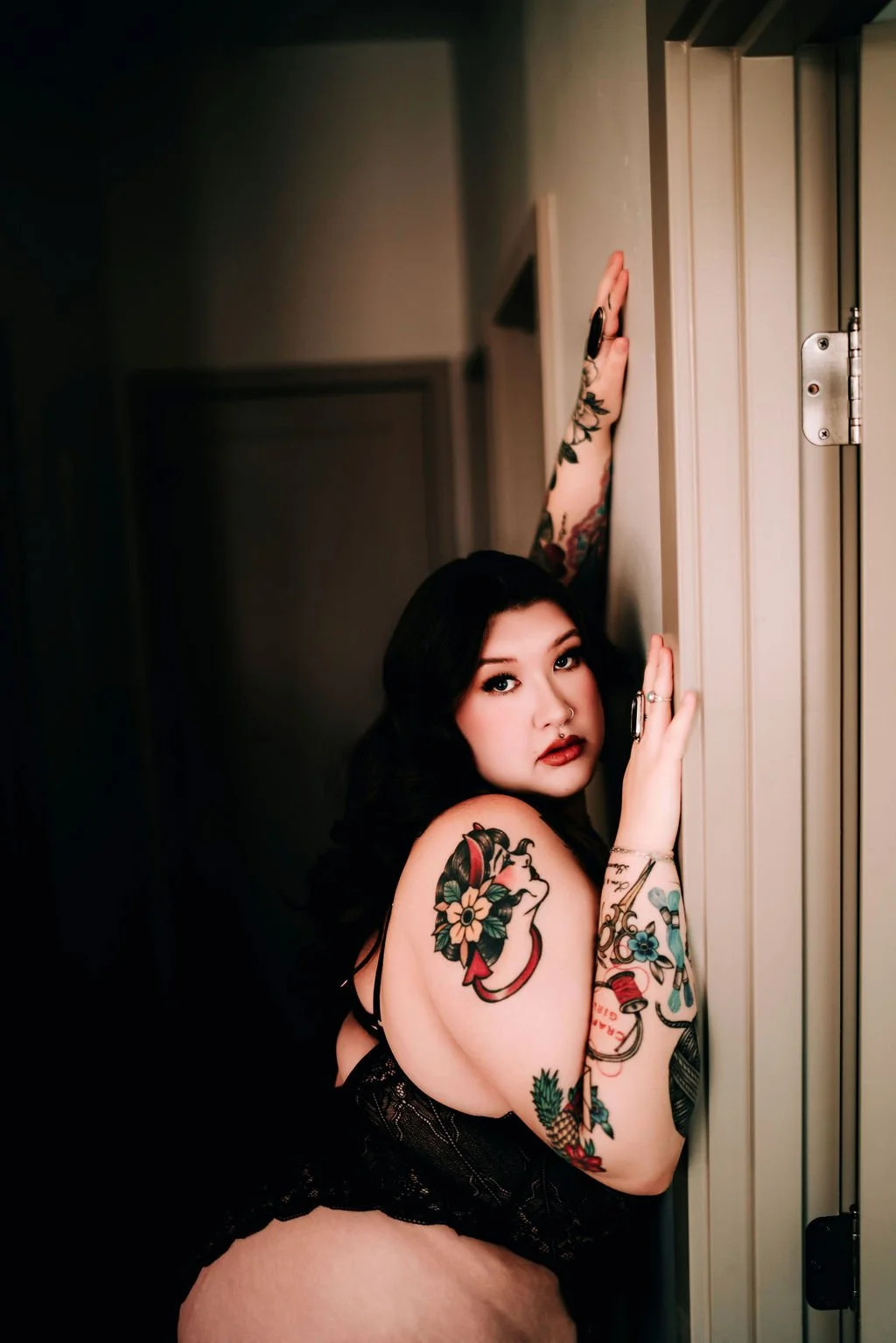 A woman with tattoos and dark hair leans against a doorframe, looking directly at the camera.