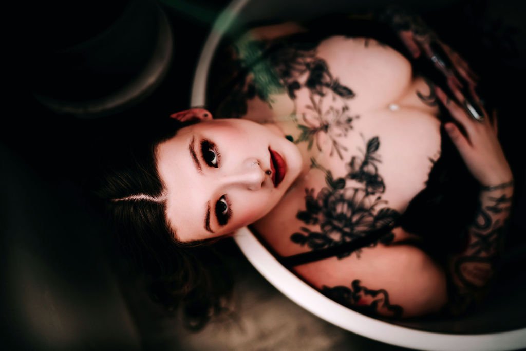 A woman with dark hair and tattoos looks up at the camera while sitting in a bathtub.