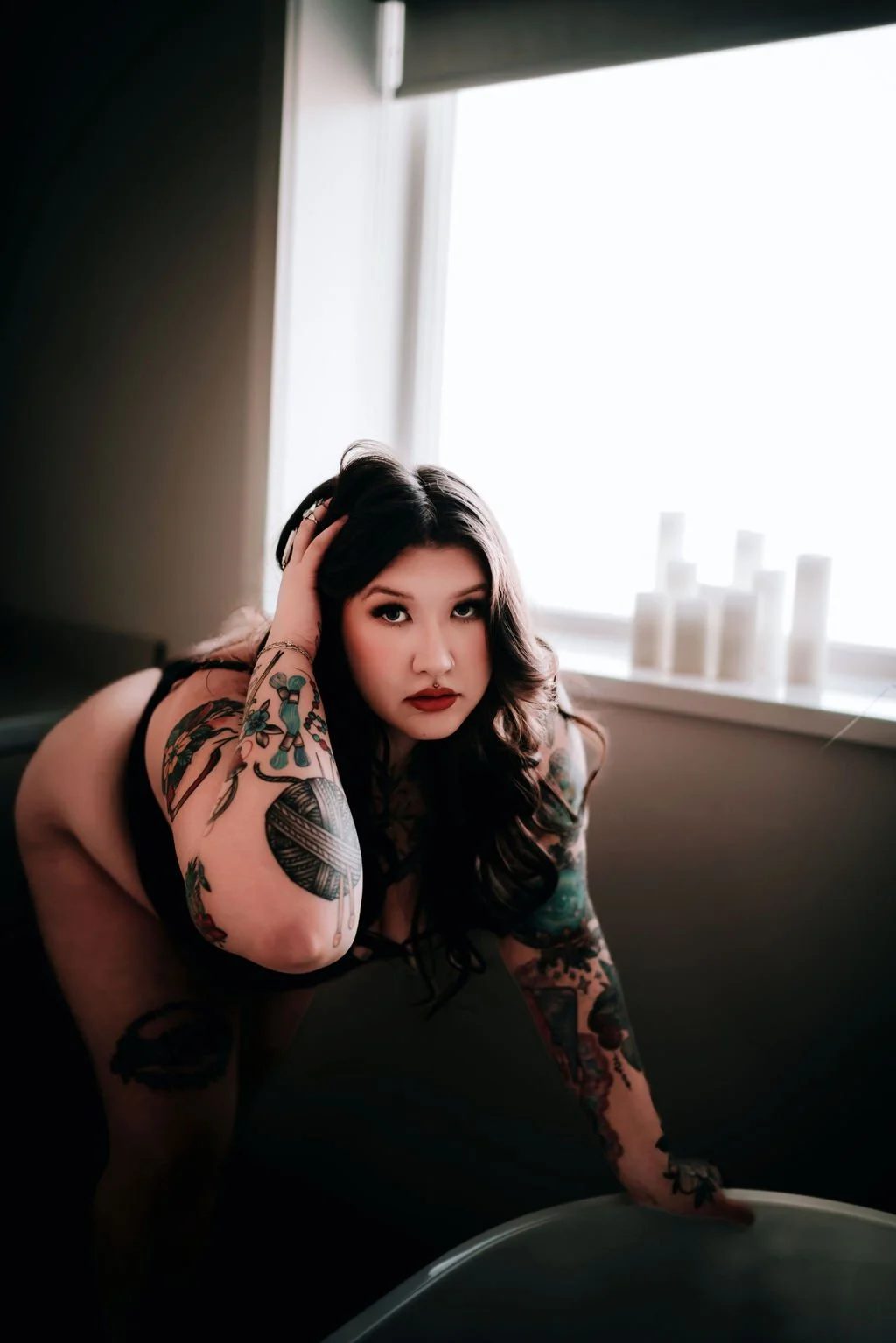A woman with long dark hair and tattoos on her arms, sitting on the floor near a window with white curtains, looking at the camera with a serious expression.