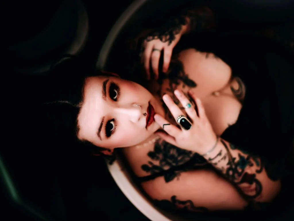 A woman with dark hair, tattoos, and jewelry lying in a car, looking up at the camera with her hand near her face.
