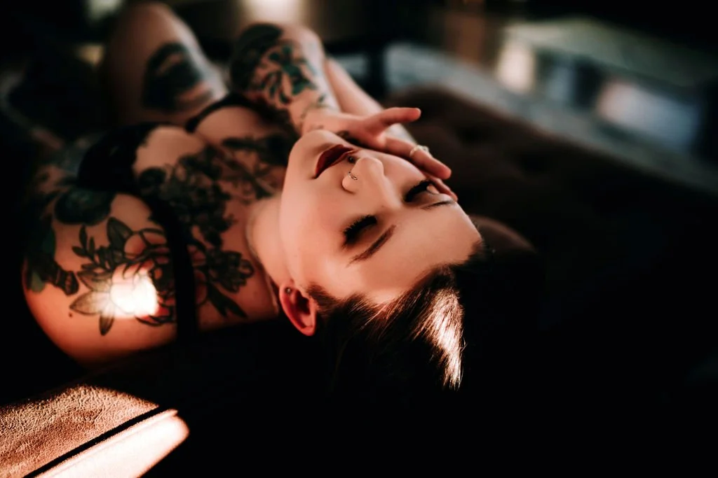 A woman with tattoos and piercings lying down with her eyes closed and her fingers resting on her face in a dimly lit room.
