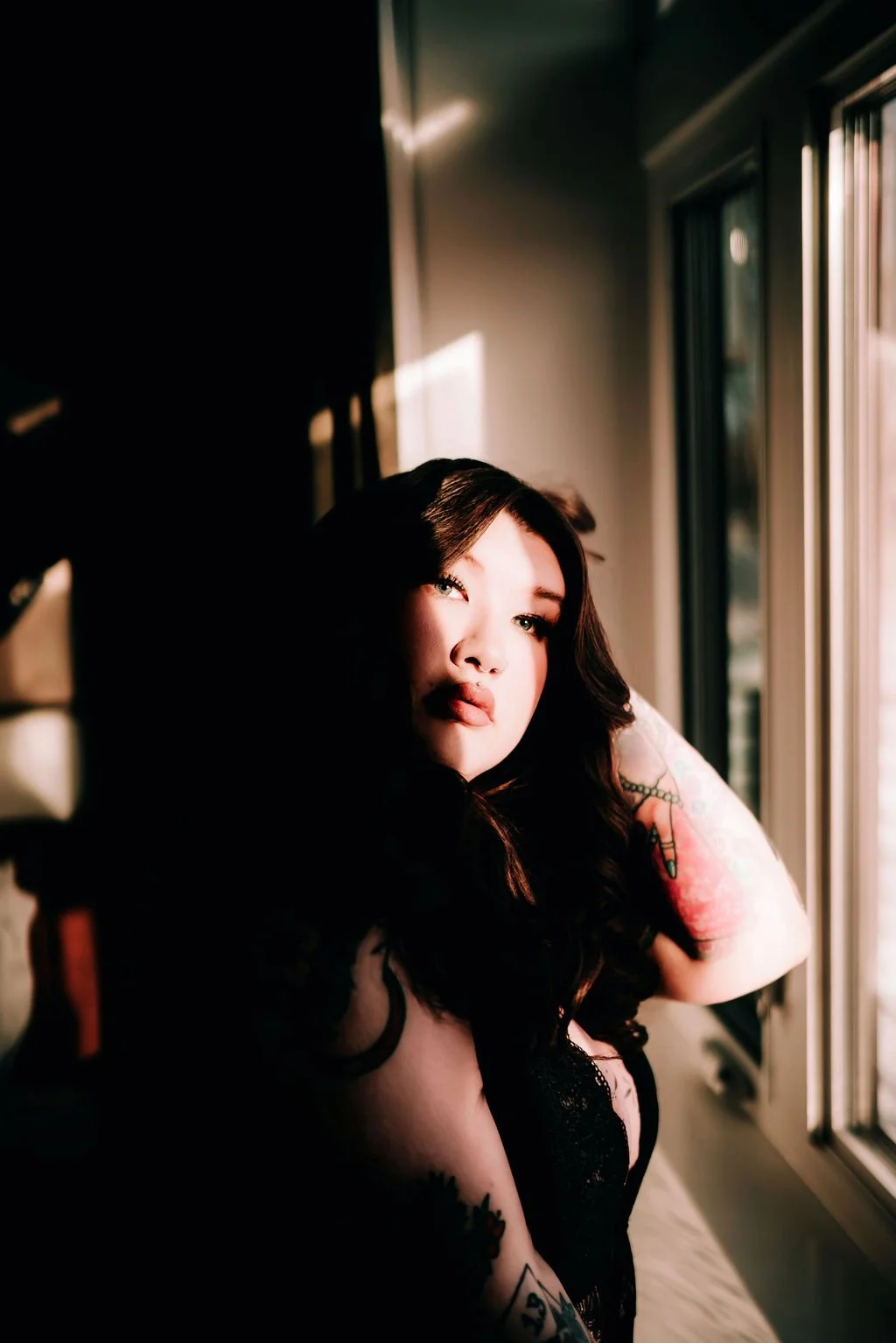 A woman with long dark hair gazing out a window in warm sunlight, wearing black lace clothing, with tattoos on her arm and shoulder.