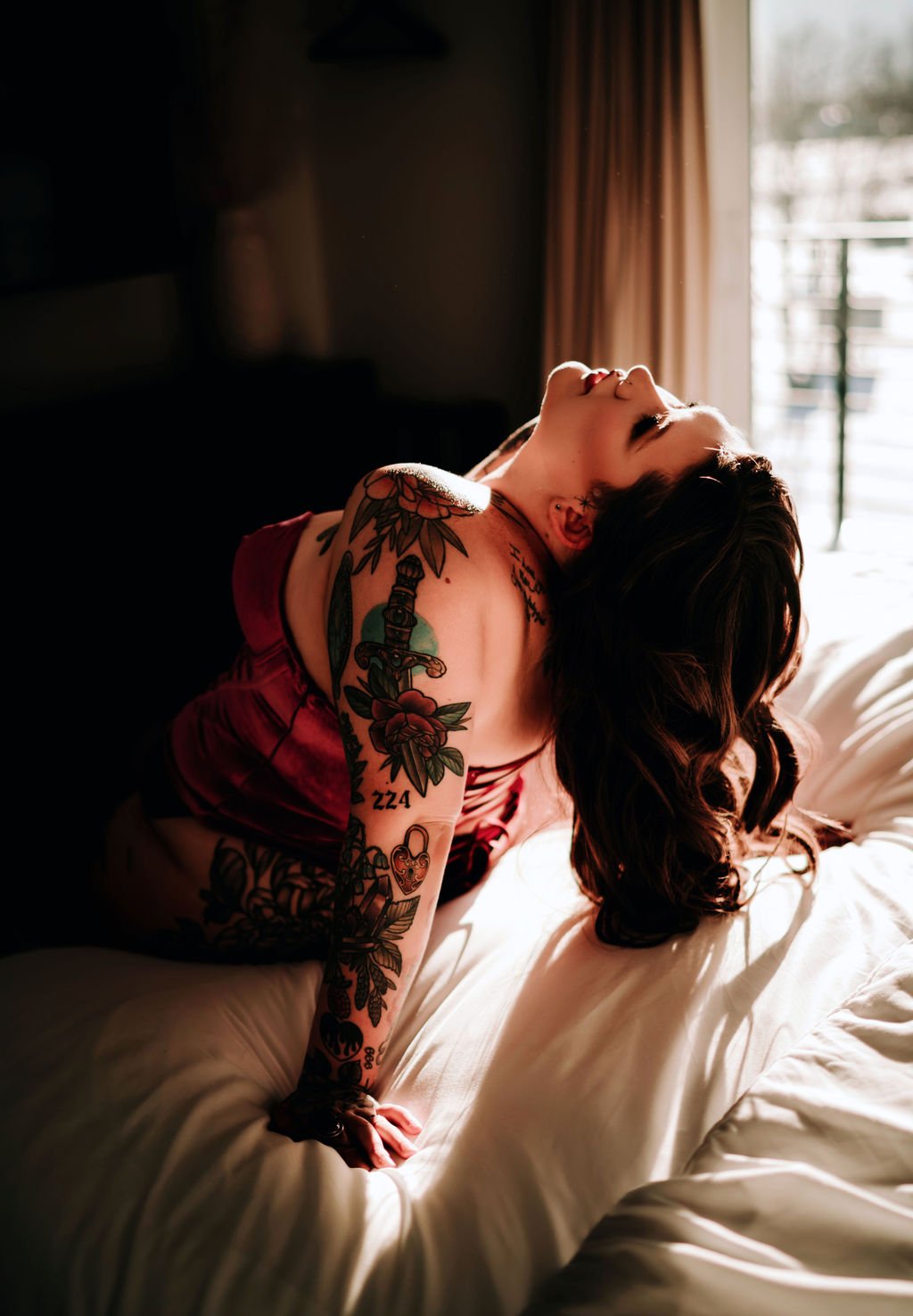 A woman with tattoos on her arm and shoulder is lying on a bed, arching her back with her head tilted back, in a sunlit room with a window and curtains.
