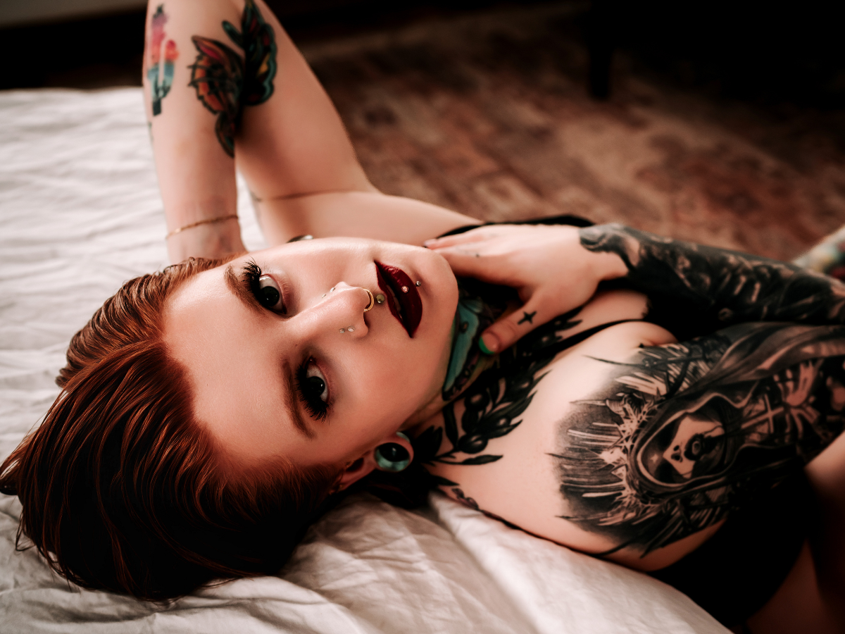 A woman with red hair lying on a bed, with tattoos, piercings, and green earrings, looking at the camera with a relaxed expression.