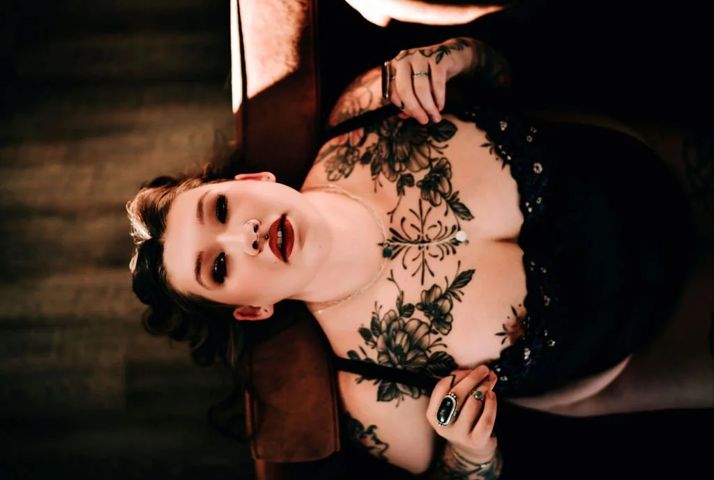 A woman with dark hair and bold makeup lying on a wooden surface, wearing a black lace dress and jewelry, with floral tattoos on her chest and arms.