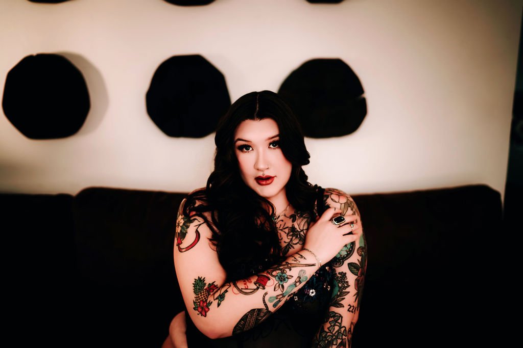 A woman with dark hair and tattoos on her arms and chest, wearing dark lipstick, sitting on a black couch with black art pieces on the wall behind her.