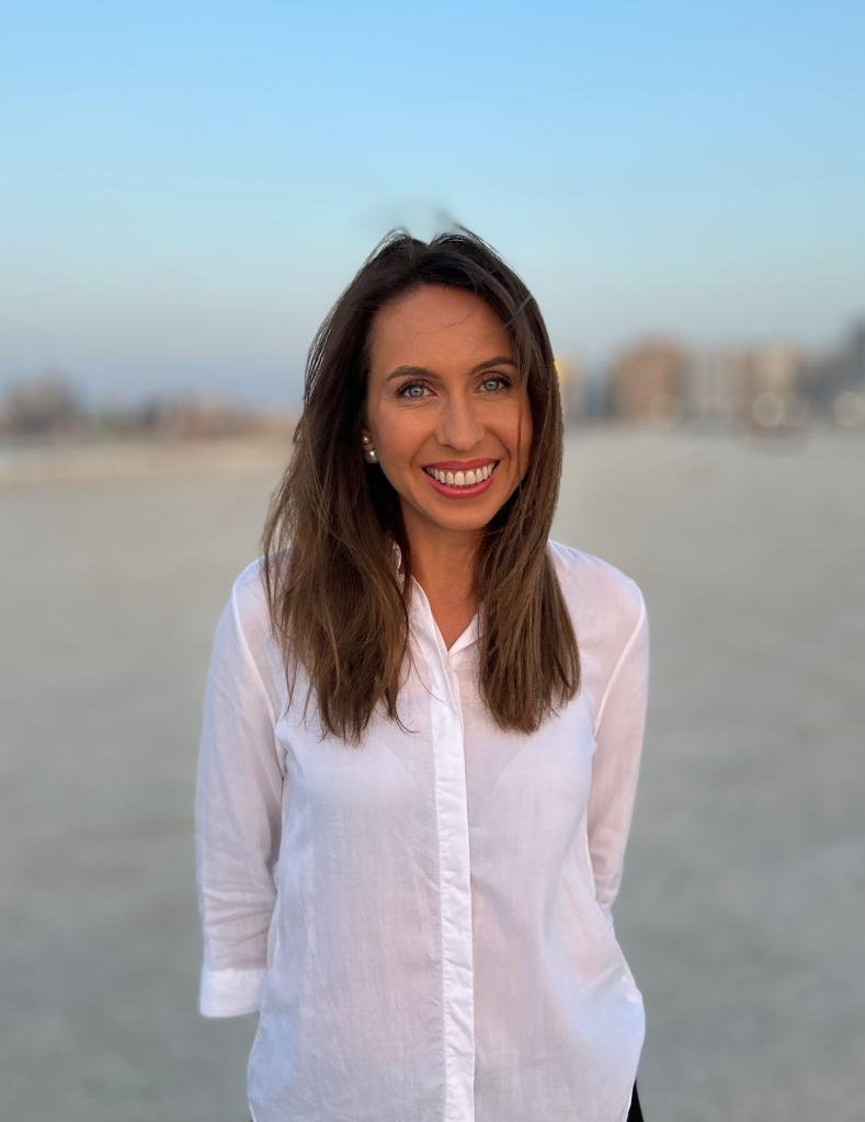 Young woman with long brown hair wearing a white long-sleeve shirt, smiling outdoors on a beach with blurry buildings and a blue sky in the background.