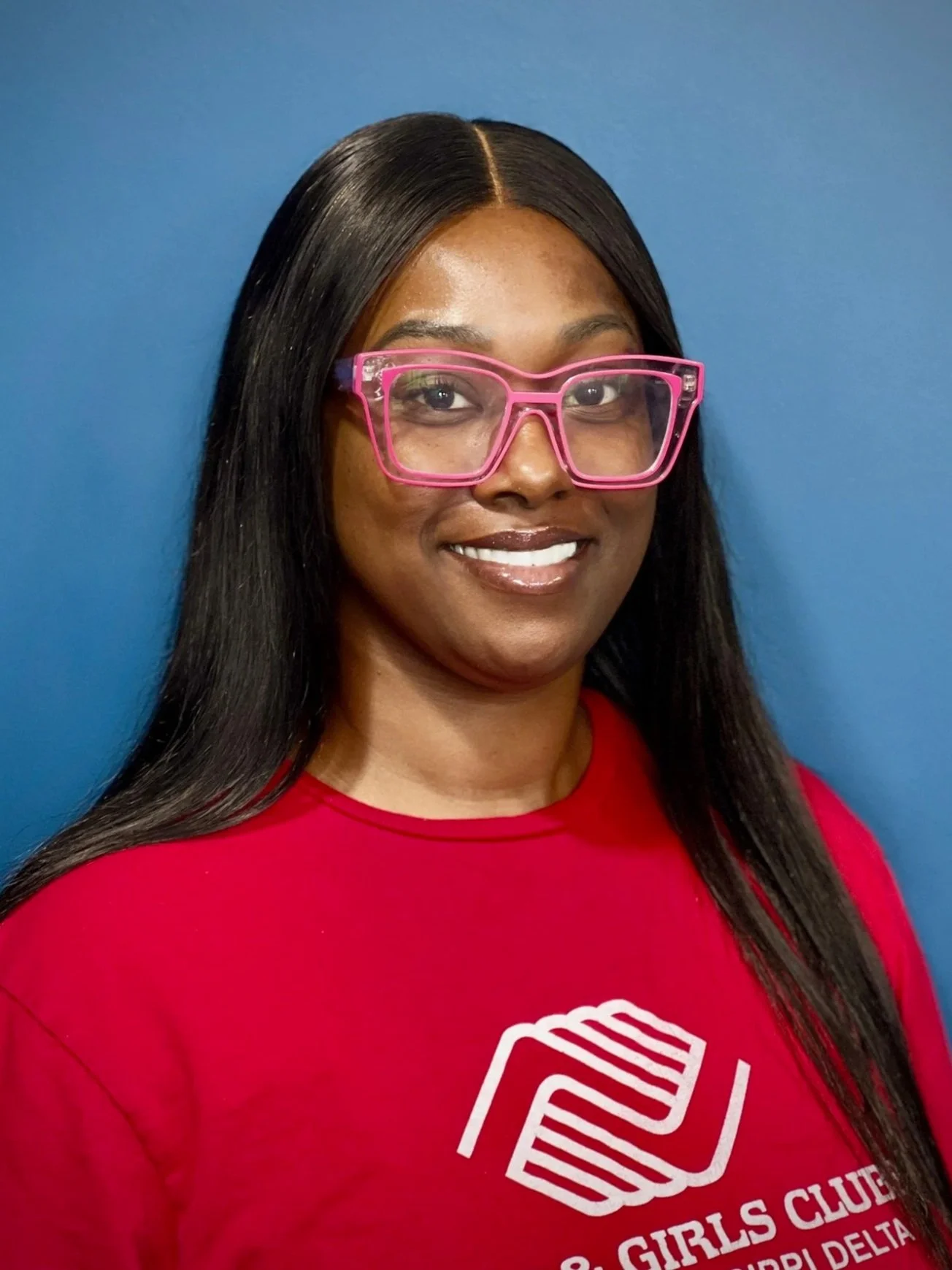 A smiling woman with long black hair and pink glasses, wearing a red shirts with a Boys & Girls Clubs logo, standing against a blue background.