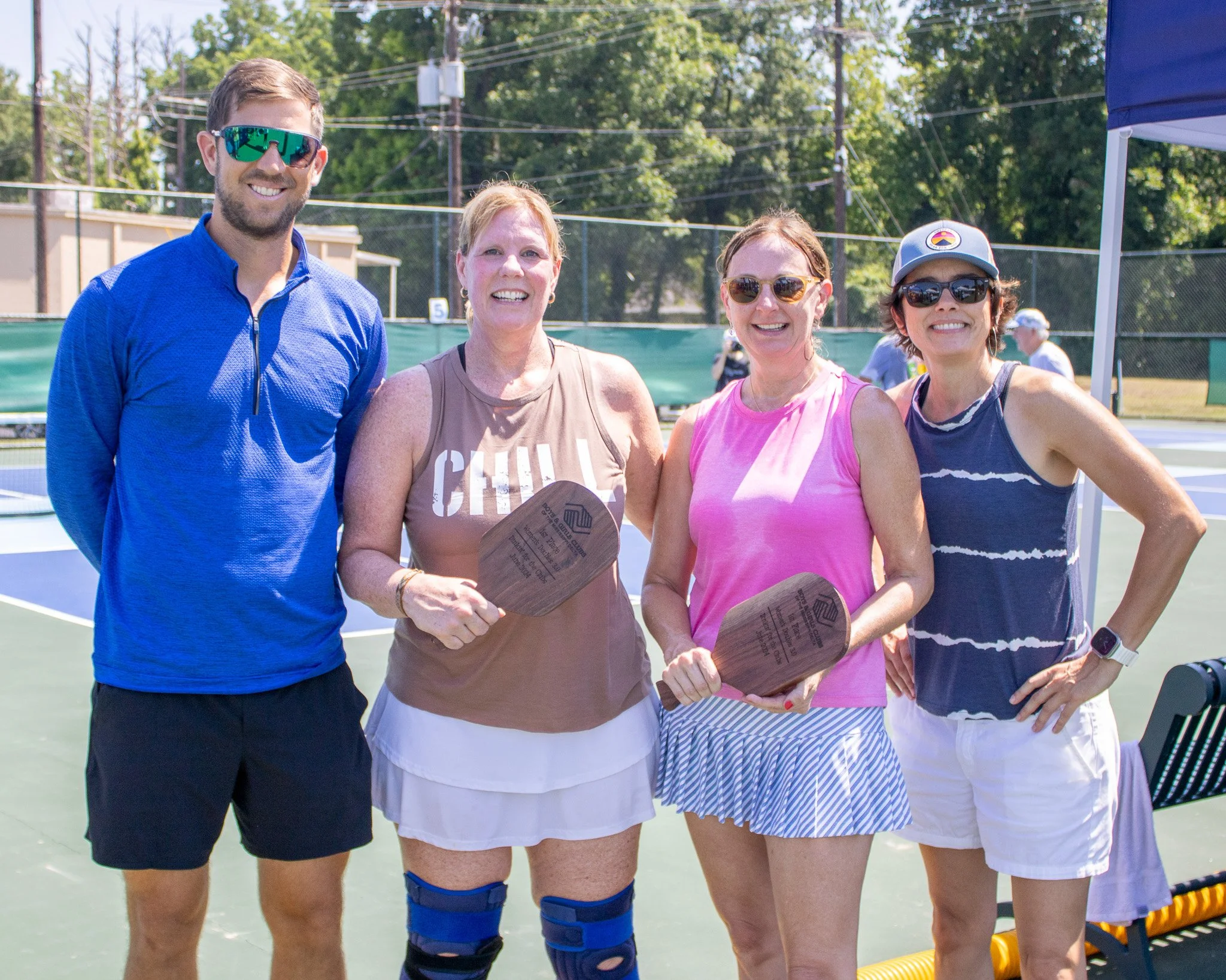 Four people standing on a tennis court, two women holding paddles, smiling after a game, with trees and a fence in the background.