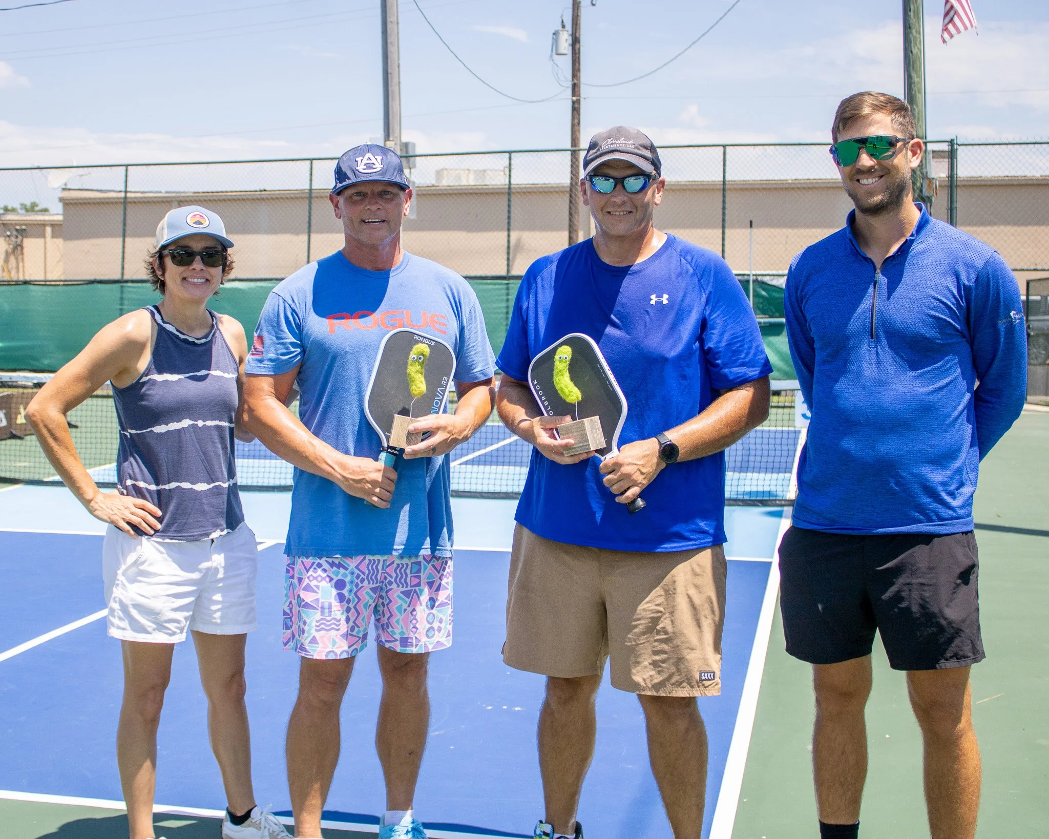 Four people standing on a tennis court. Two men are holding paddle awards, and the woman on the left has her hand on her hip. All are dressed in athletic clothing, wearing sunglasses, and smiling. The background shows a chain-link fence and a bright, partly cloudy sky.