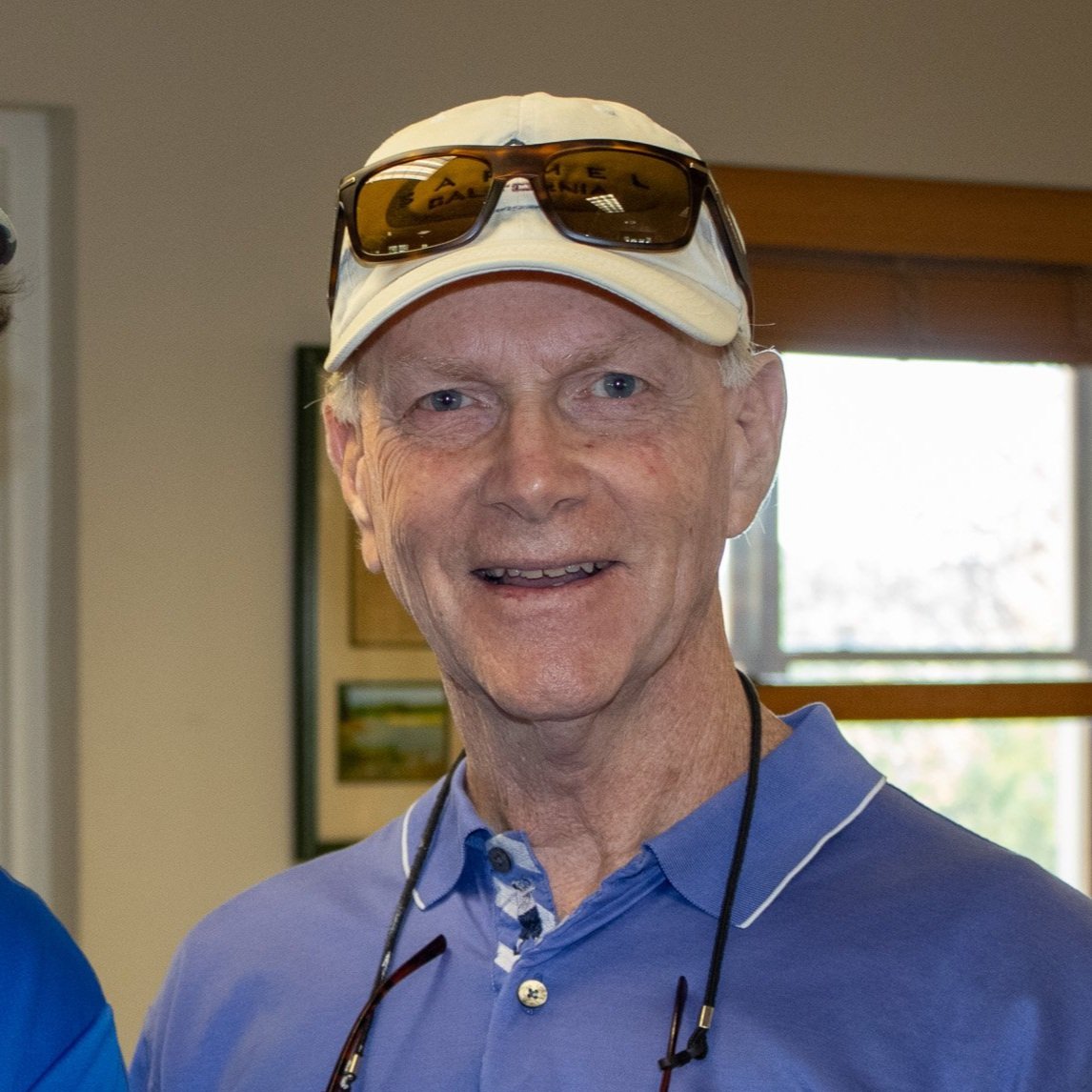 A smiling man wearing a white baseball cap, sunglasses on the cap, and a purple polo shirt, standing indoors near a window.