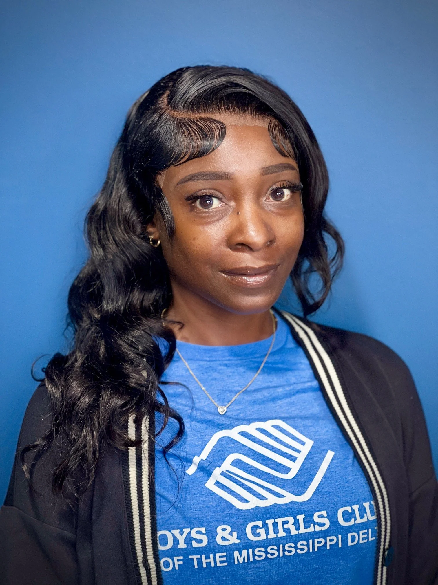 Portrait of a woman wearing a blue T-shirt with the Boys & Girls Club logo, a black jacket, and a necklace, standing against a blue background.