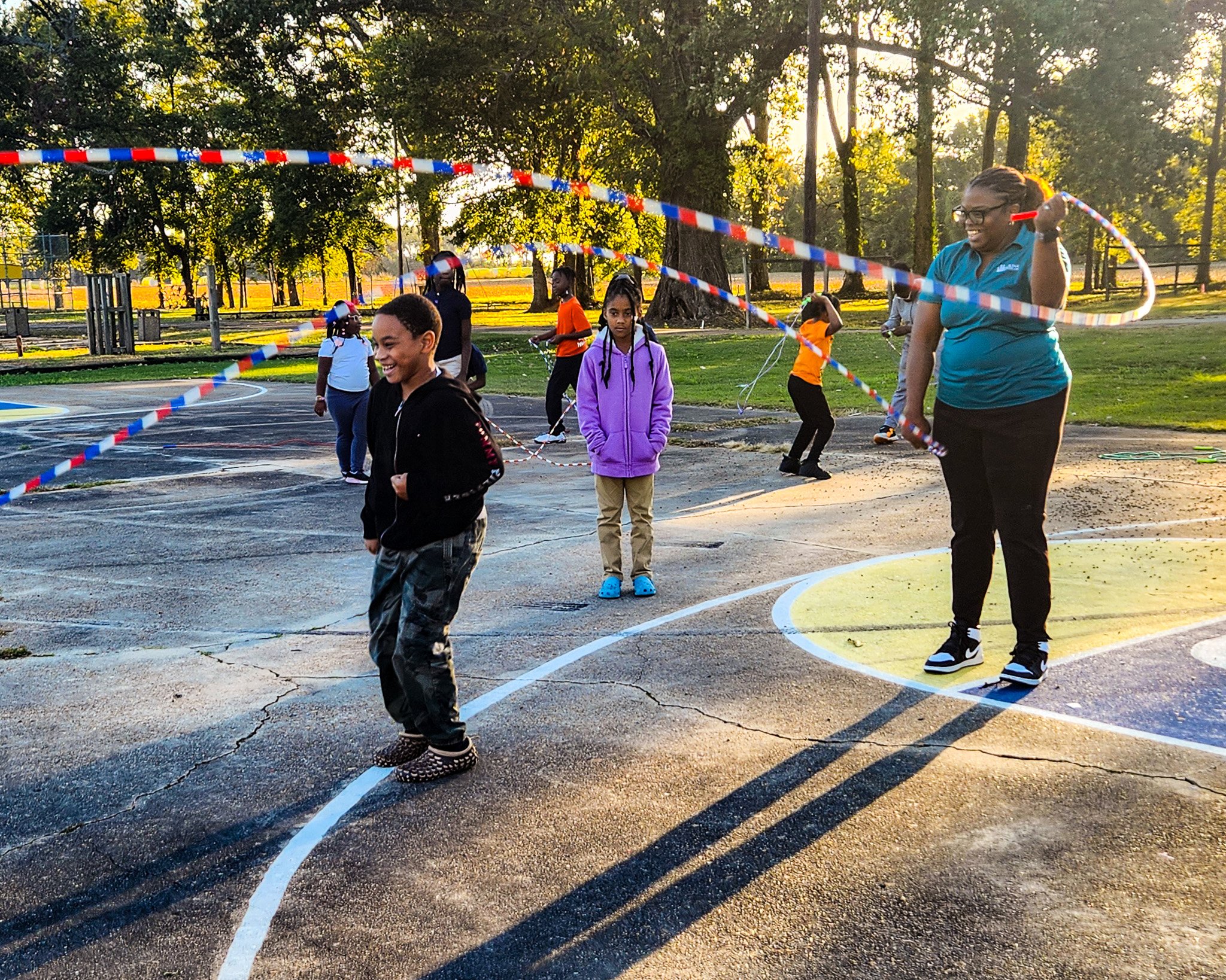 Children and an adult playing with a jump rope on a basketball court in a park during sunset.